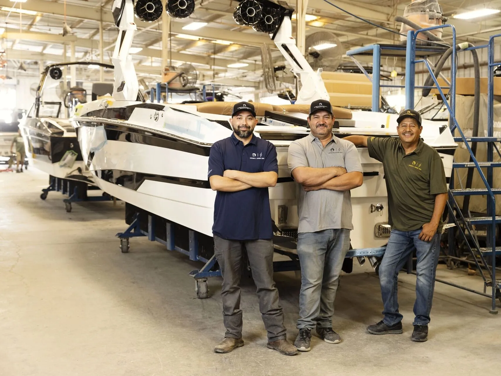 Three men in work uniforms stand in front of a large Centurion boat inside a spacious, well-lit industrial workshop, ready to begin a factory tour.