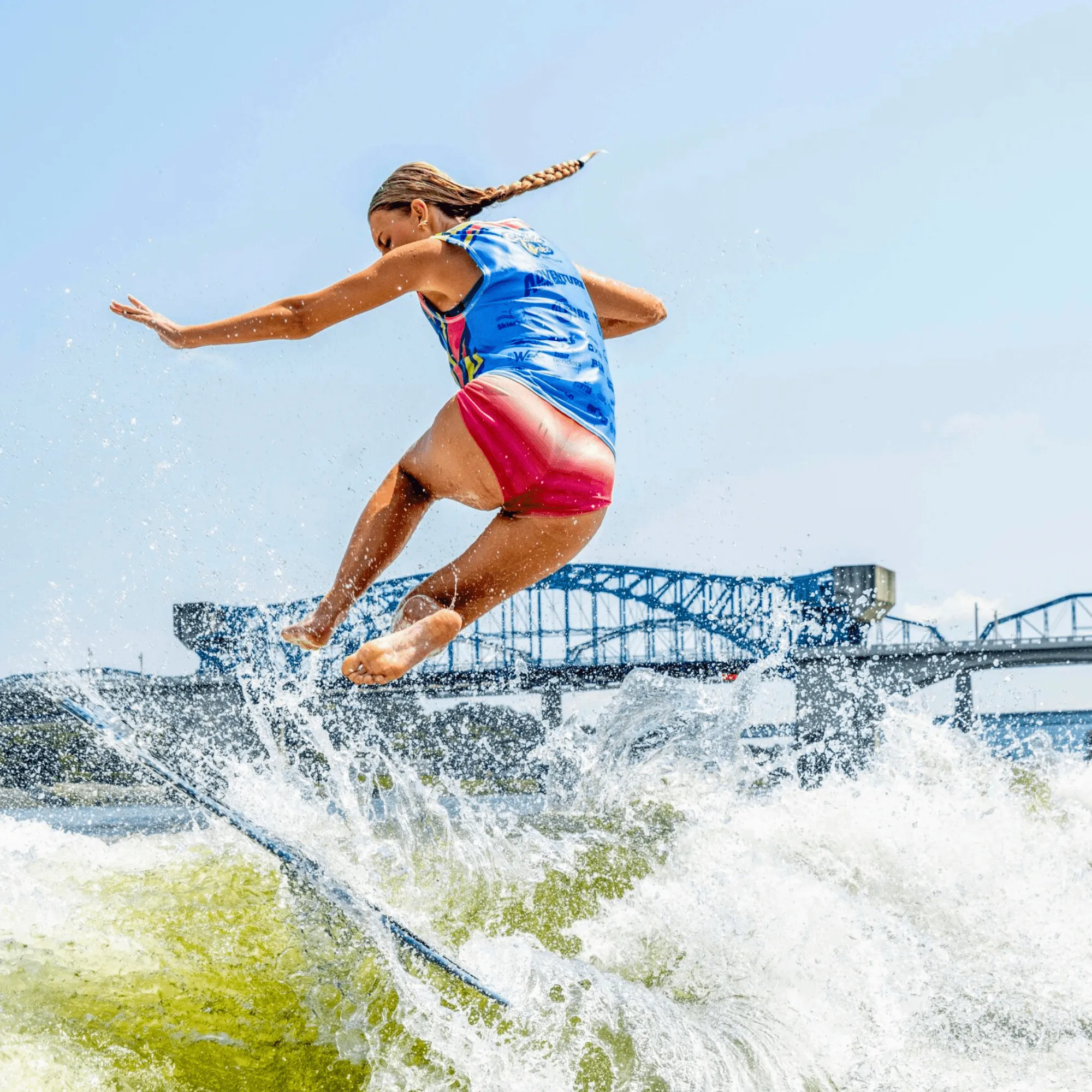 Ava Stewart, in a blue vest and red shorts, is captured mid-air while surfing on a wave, with a blue bridge structure visible in the background.