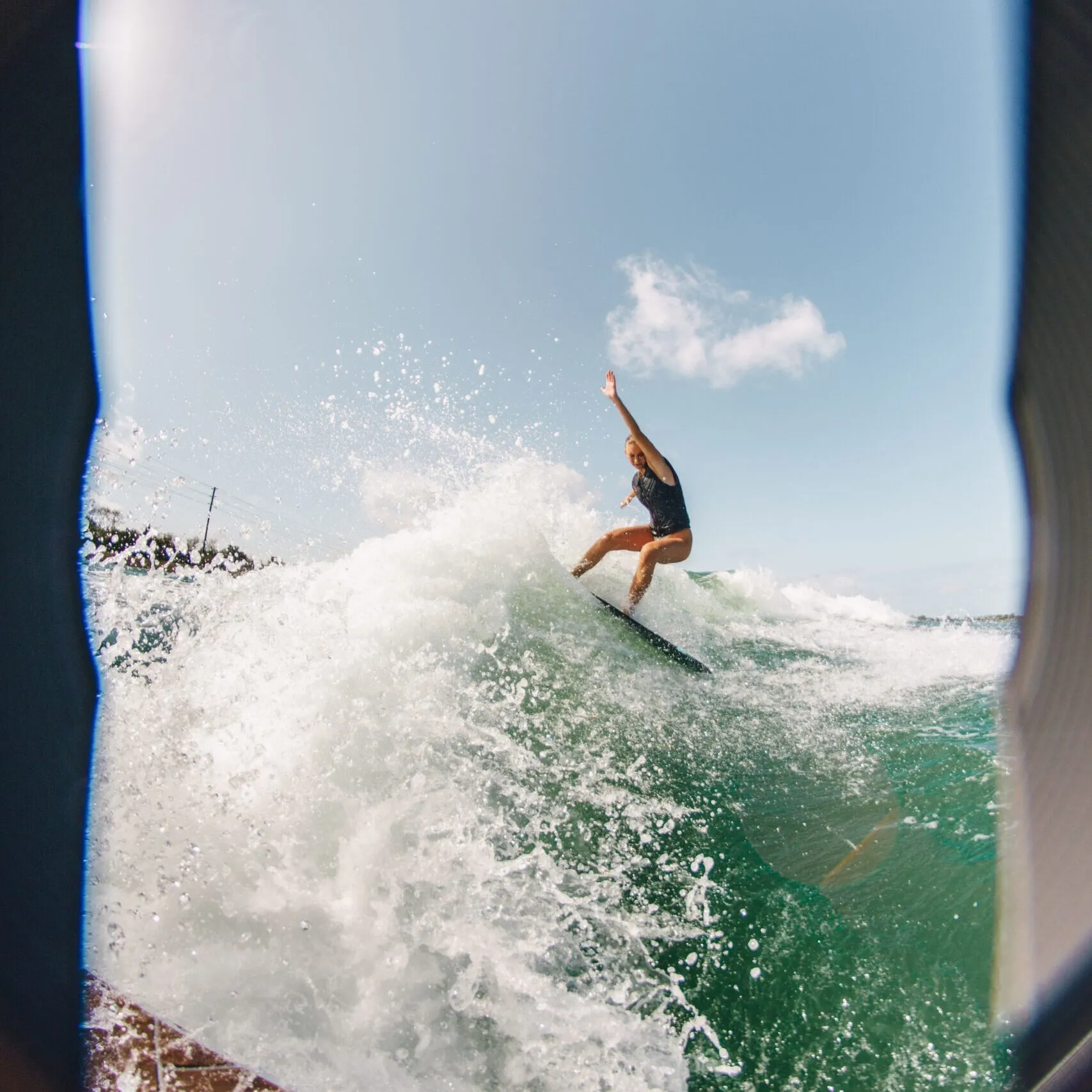 Ava Stewart rides a wave on a surfboard, captured from a low angle with water splashing and a bright blue sky in the background.