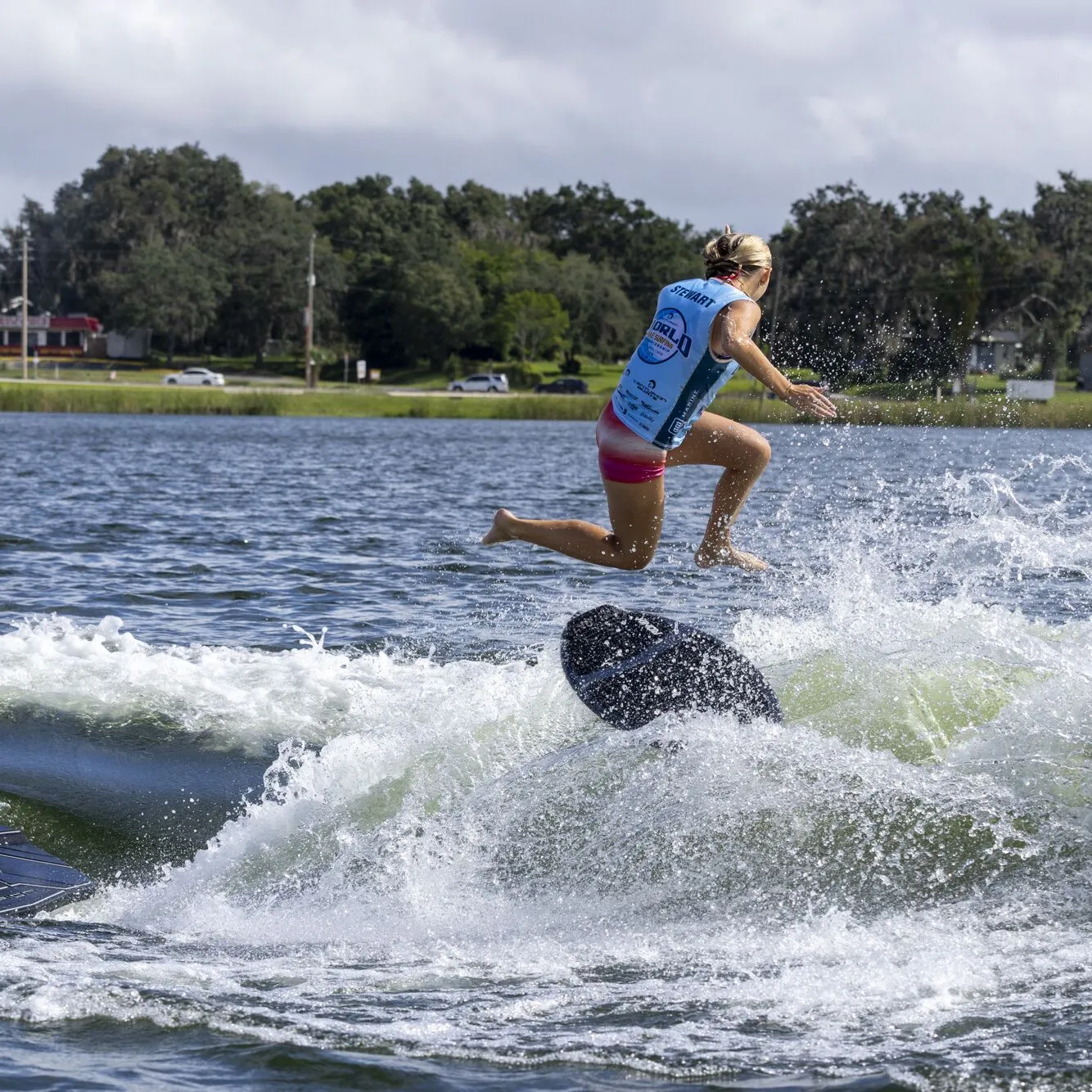 Ava Stewart, wearing a blue vest and pink shorts, falls off a wakesurf board and splashes into the lake behind a boat.