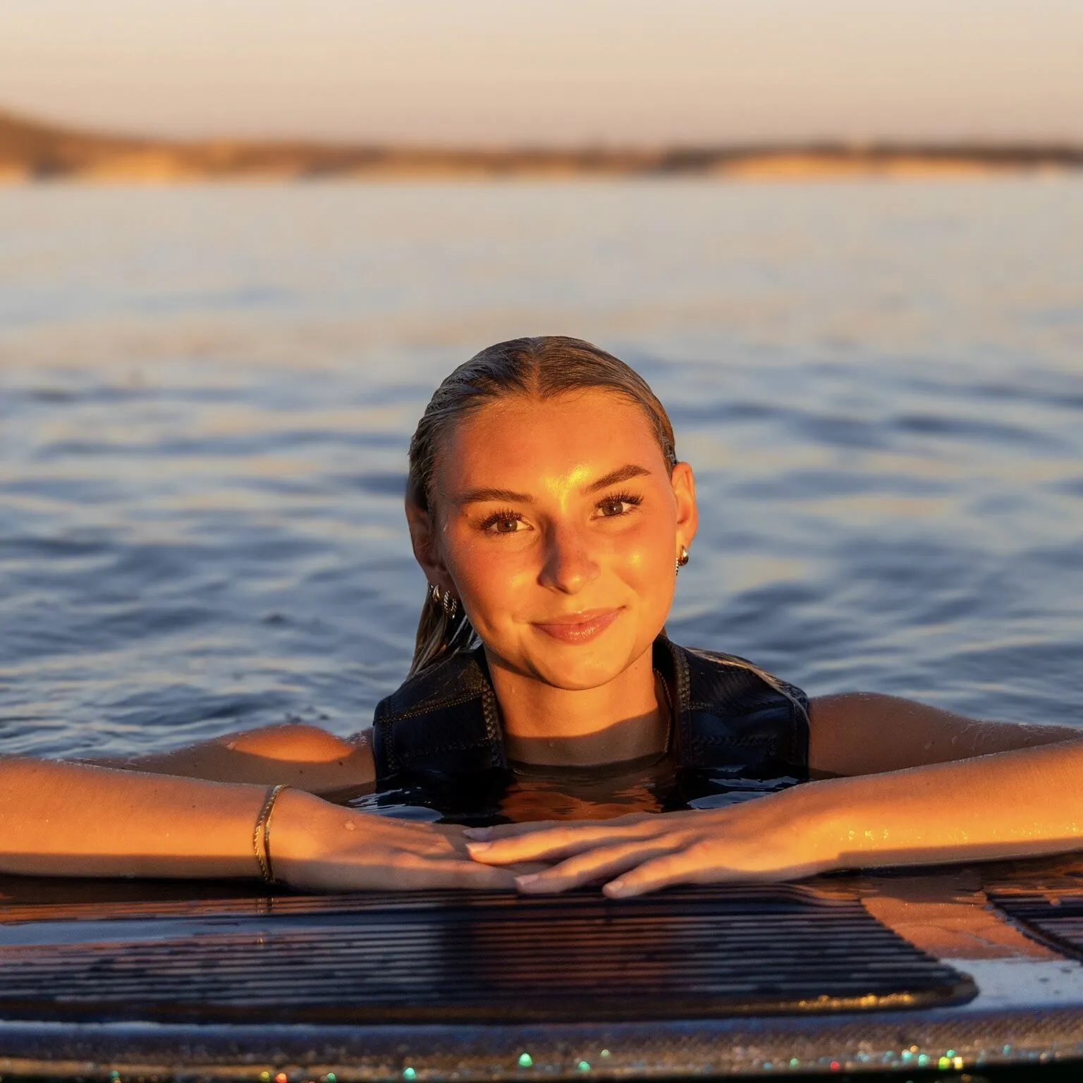 Ava Stewart, wearing a life vest, rests her arms on a surfboard in calm water at sunset, with golden light reflecting on her face.