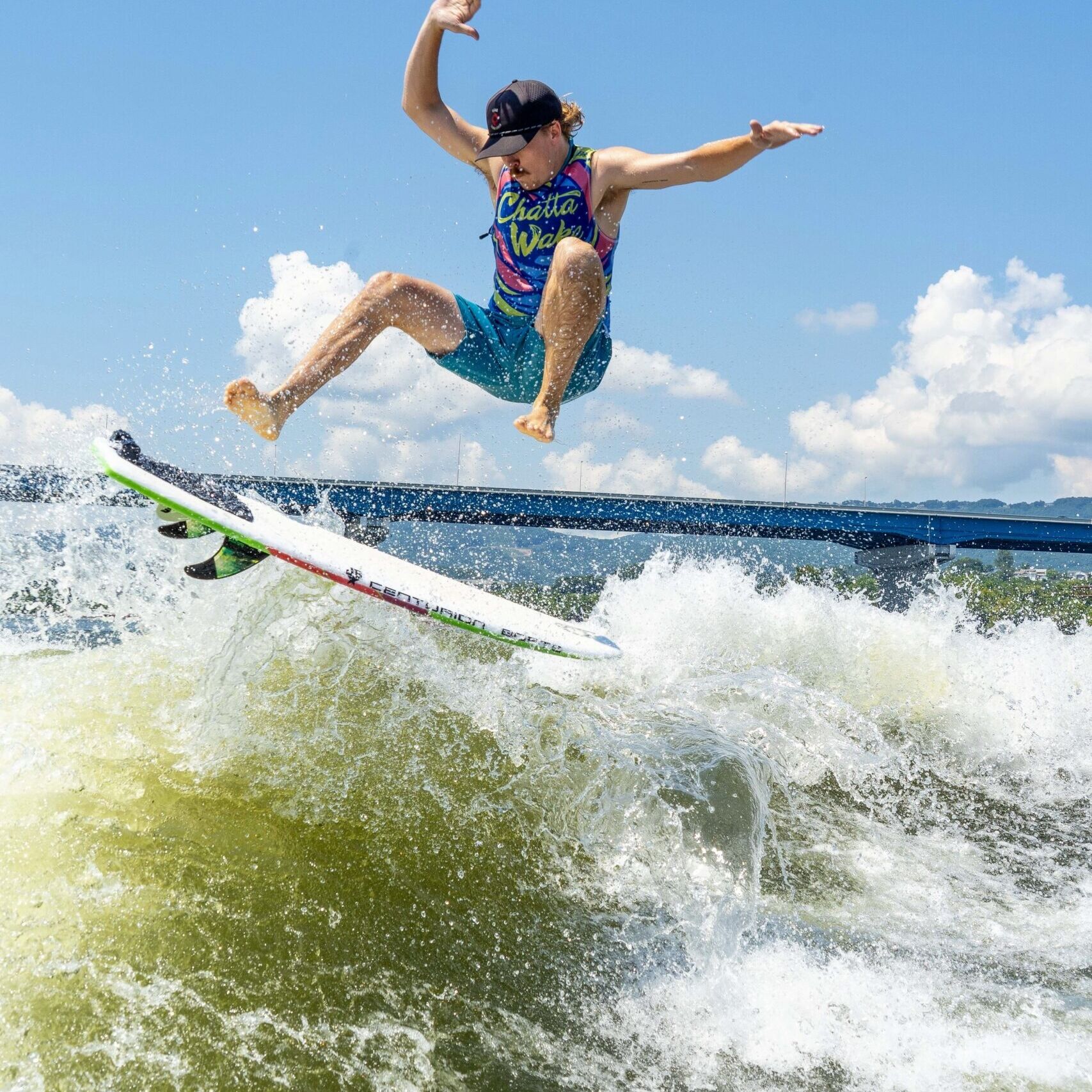 A person in colorful clothes and a cap is performing an aerial trick above the water on a surfboard, with waves splashing beneath and a blue sky in the background.