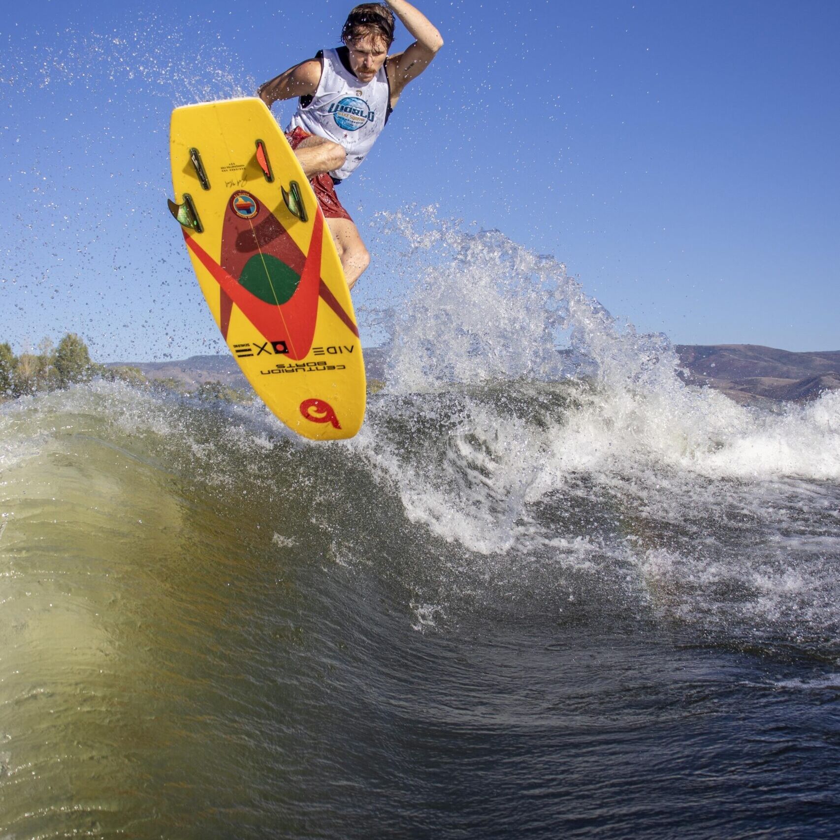 A person wearing a competition jersey rides a yellow wakeboard over a wave, airborne above the water with a clear blue sky in the background.