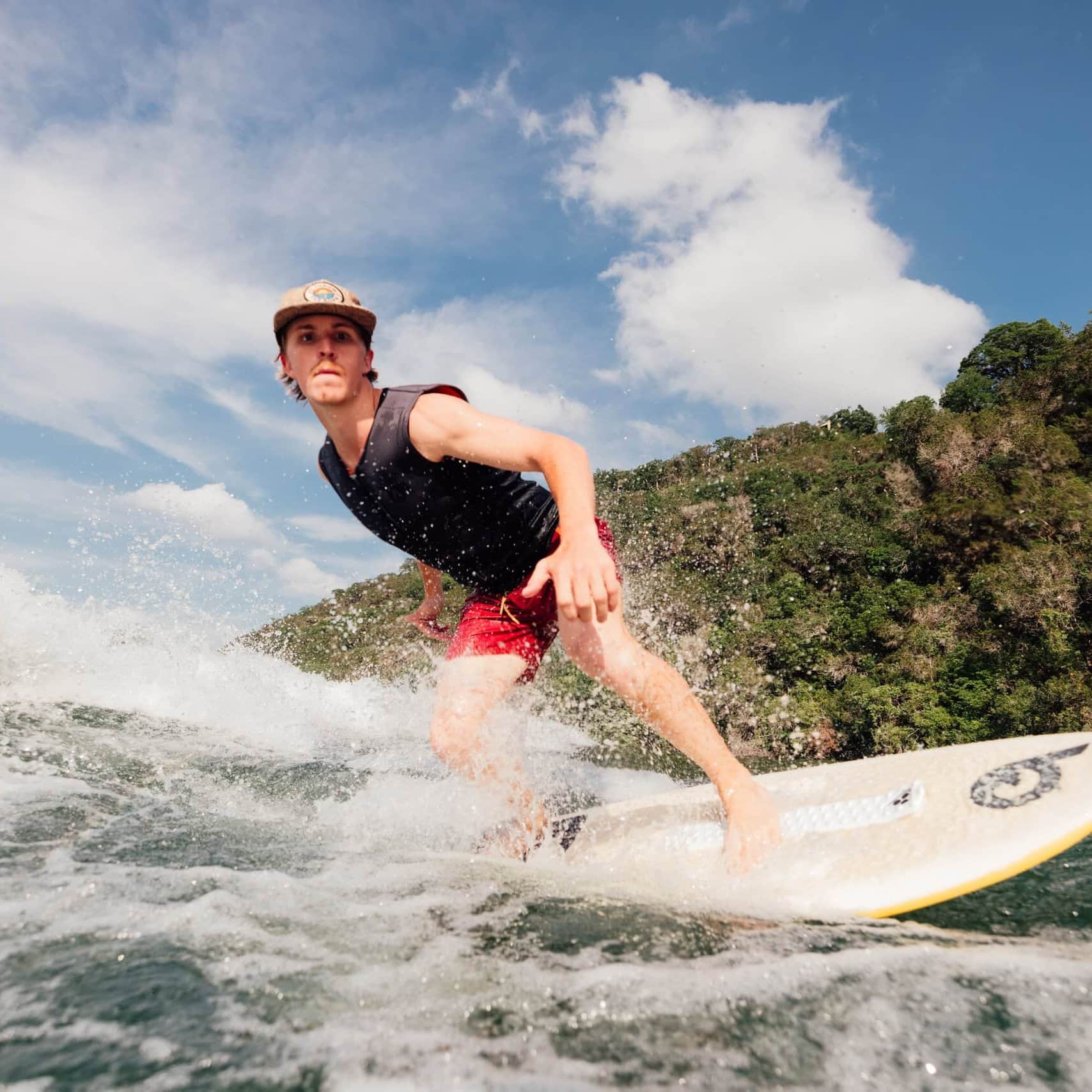 A person in a black vest and red shorts rides a wave on a surfboard near a green, tree-covered shoreline under a partly cloudy sky.