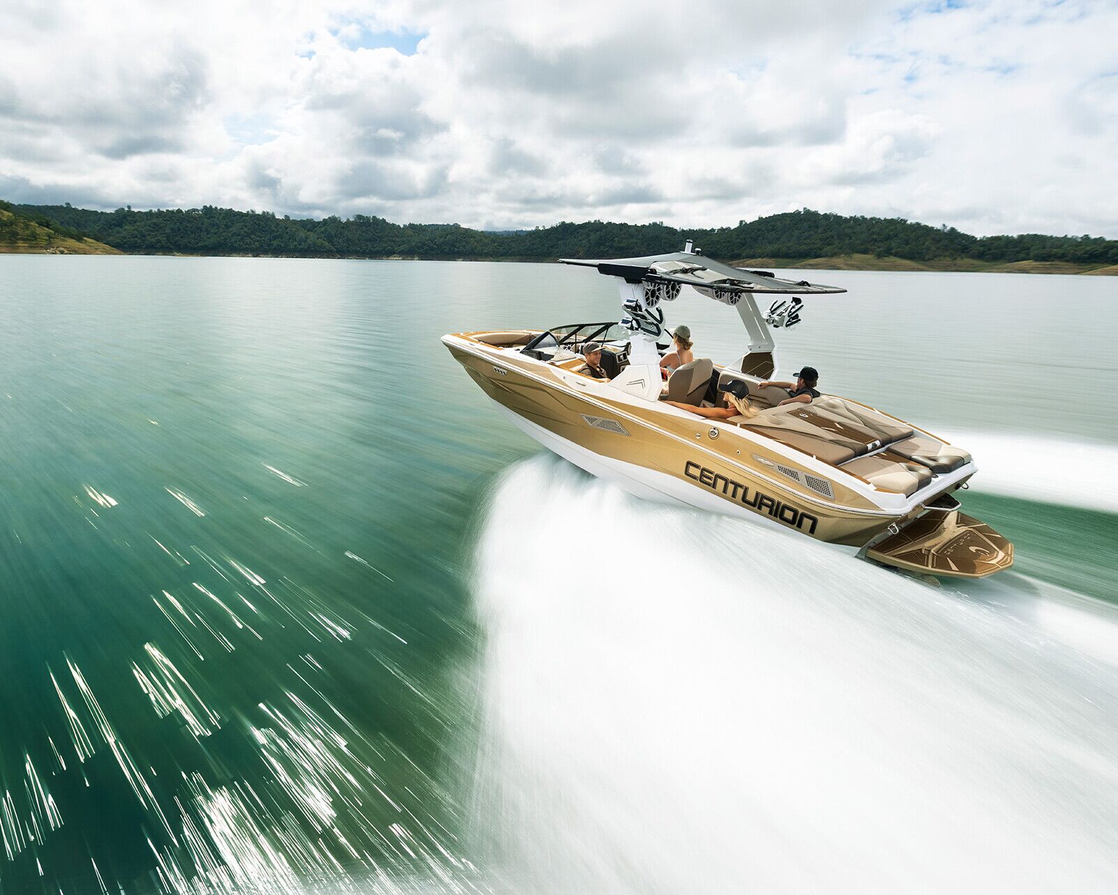 A gold Centurion Nv213 wake boat with several people onboard moves quickly across a calm lake, leaving white wakes behind, with forested hills in the background.