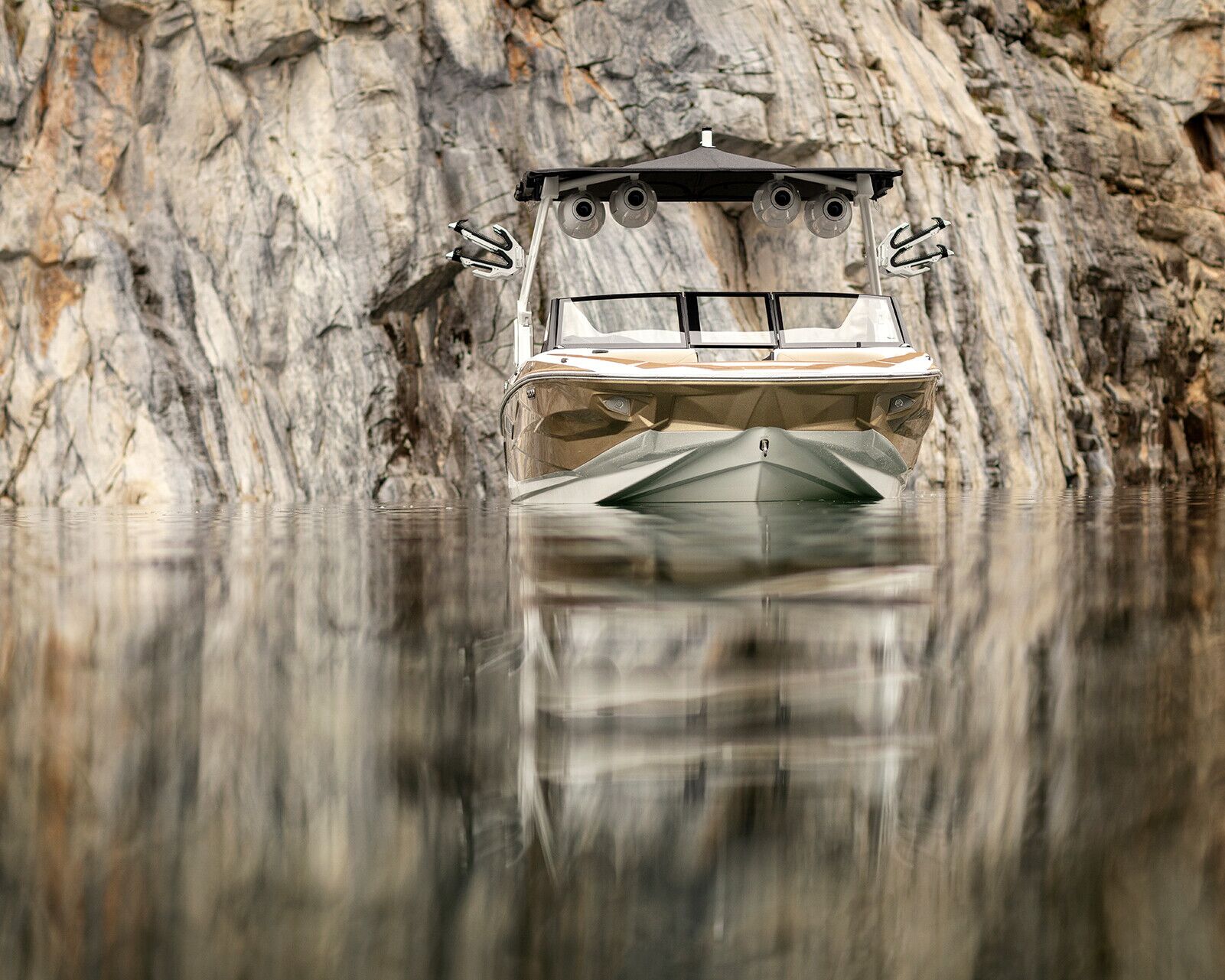A modern Centurion Nv213 surf boat is stationary on calm water near a steep, rocky cliff. The water reflects the sleek boat and the rugged rock face.