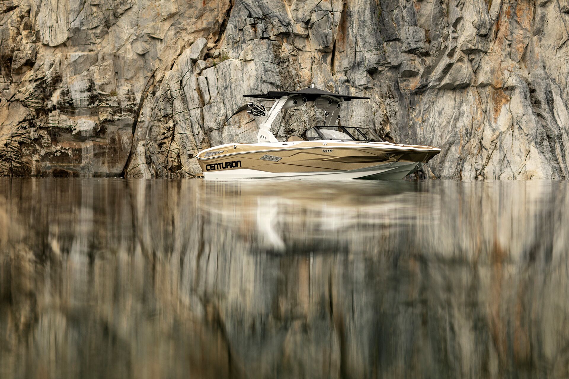 A white and gold Centurion Nv213 floats on calm water in front of a rocky cliff, with the boat and cliff perfectly reflected in the water.