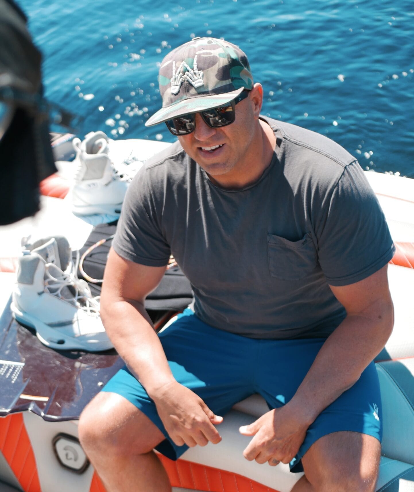 A man wearing a camouflage hat, sunglasses, grey t-shirt, and blue shorts sits on a boat near water sports gear with water in the background.