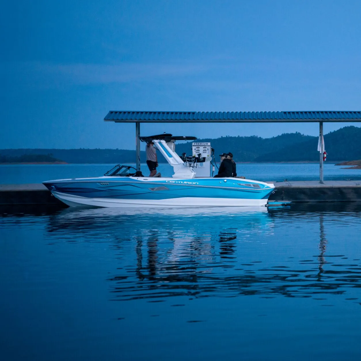 A blue and white speedboat is docked beside a covered fuel station on a calm lake at dusk, with hills visible in the background.