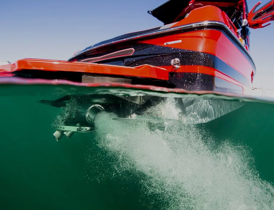 A red boat with a Centurion Opti-V Hull is partially submerged in water, bubbles trailing from its propeller, as seen from a split above-and-below water perspective.