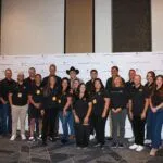 A group of people poses together in front of a branded step-and-repeat banner at a dealership event. Most are wearing matching black shirts and event lanyards.