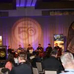A panel of six people speaks on stage at a conference in front of an audience, with banners and screens displaying agricultural images and a "50" logo in the background.