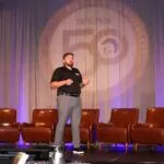 A man stands on stage in front of five empty brown chairs, speaking during a presentation with agricultural images and a 50th-anniversary logo projected behind him.