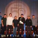 Eight men stand in a row on stage, one holding a plaque. A large "50" anniversary logo is projected on the curtain behind them.