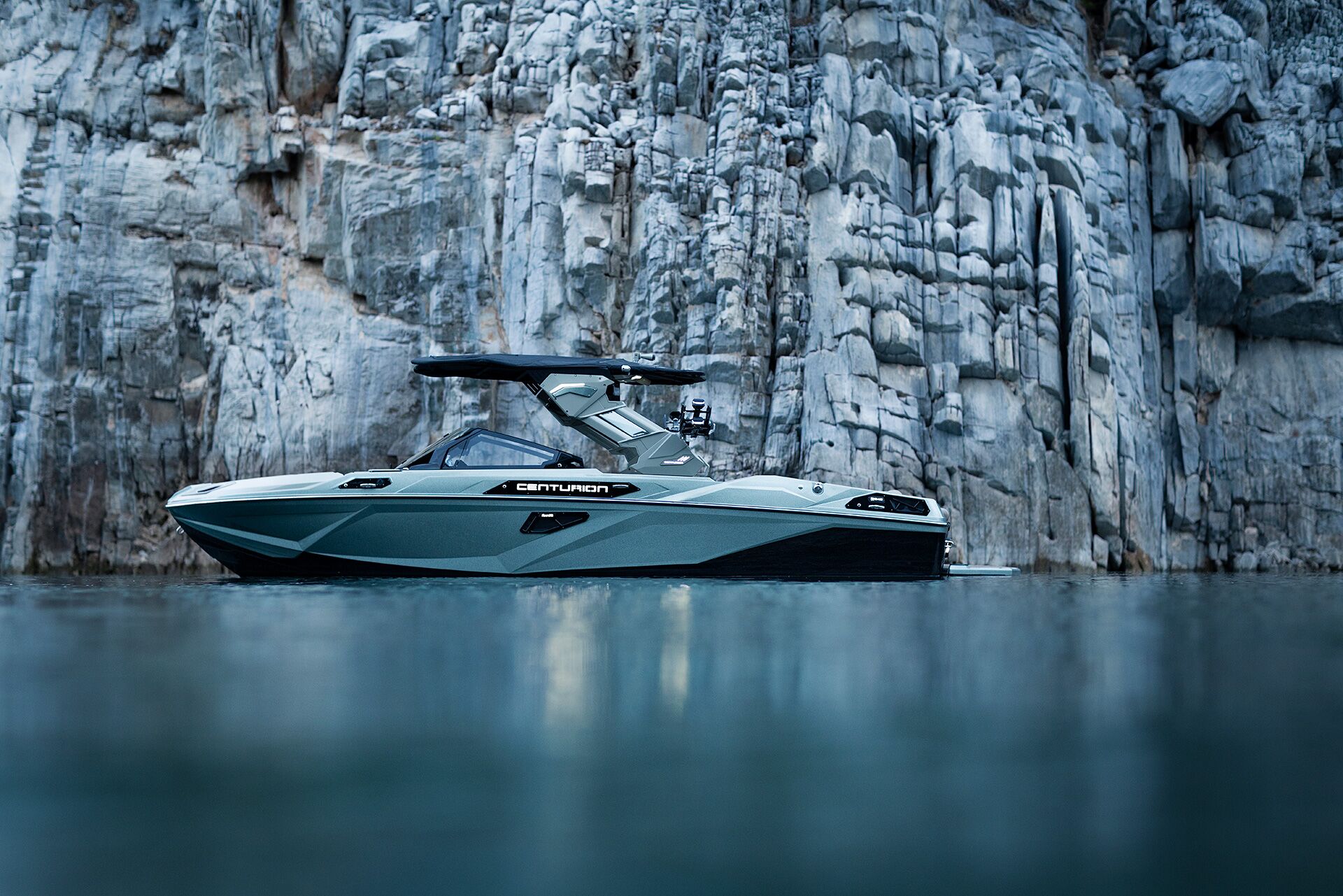 A modern Centurion boat floats on calm water near a steep, rocky cliff.
