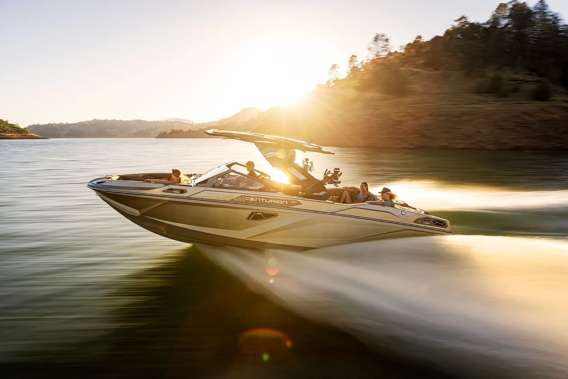 A speedboat with several passengers moves quickly across a lake at sunset, creating a wake, with a rocky shoreline and trees in the background.