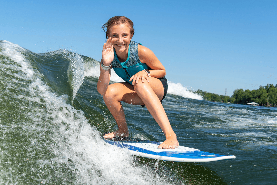 A young woman in a teal vest surfs on a wave, crouching on her board and smiling while waving with her right hand. The sky is clear and blue.