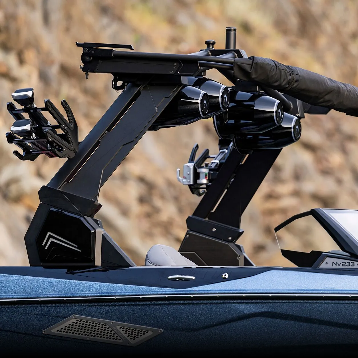 Close-up of a Centurion Towers wakeboard boat's tower with attached wakeboard racks, loudspeakers, and a folded black canopy, set against a blurred rocky background.