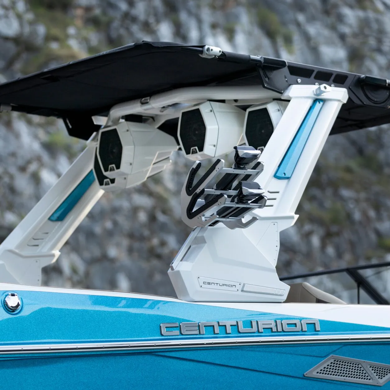 Close-up of a blue Centurion boat featuring a sleek white Centurion Towers wakeboard tower with racks, speakers, and a black sunshade, set against rocky terrain in the background.