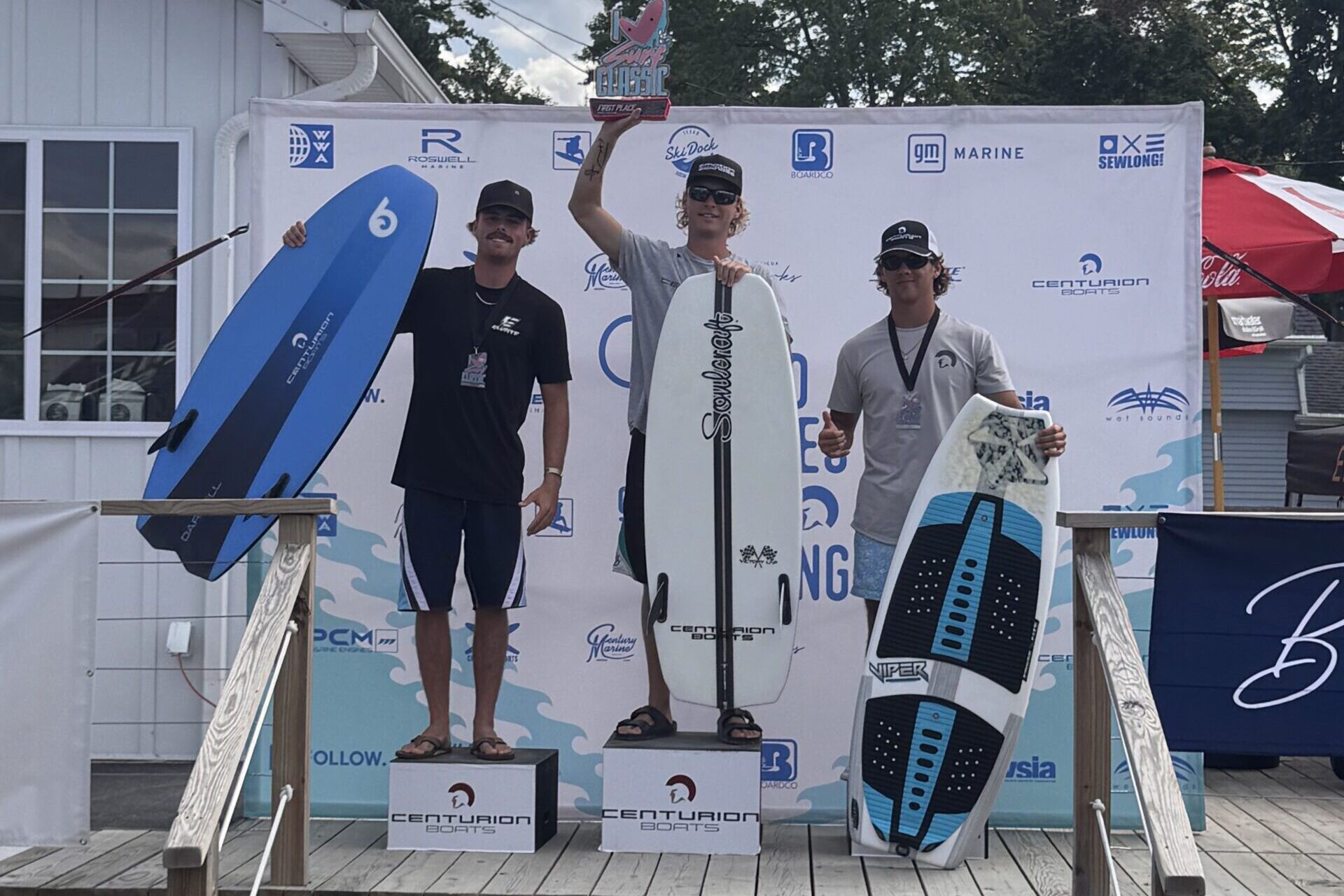 Three surfers stand on a podium holding boards, posing for a photo at a surfing competition award ceremony outdoors.