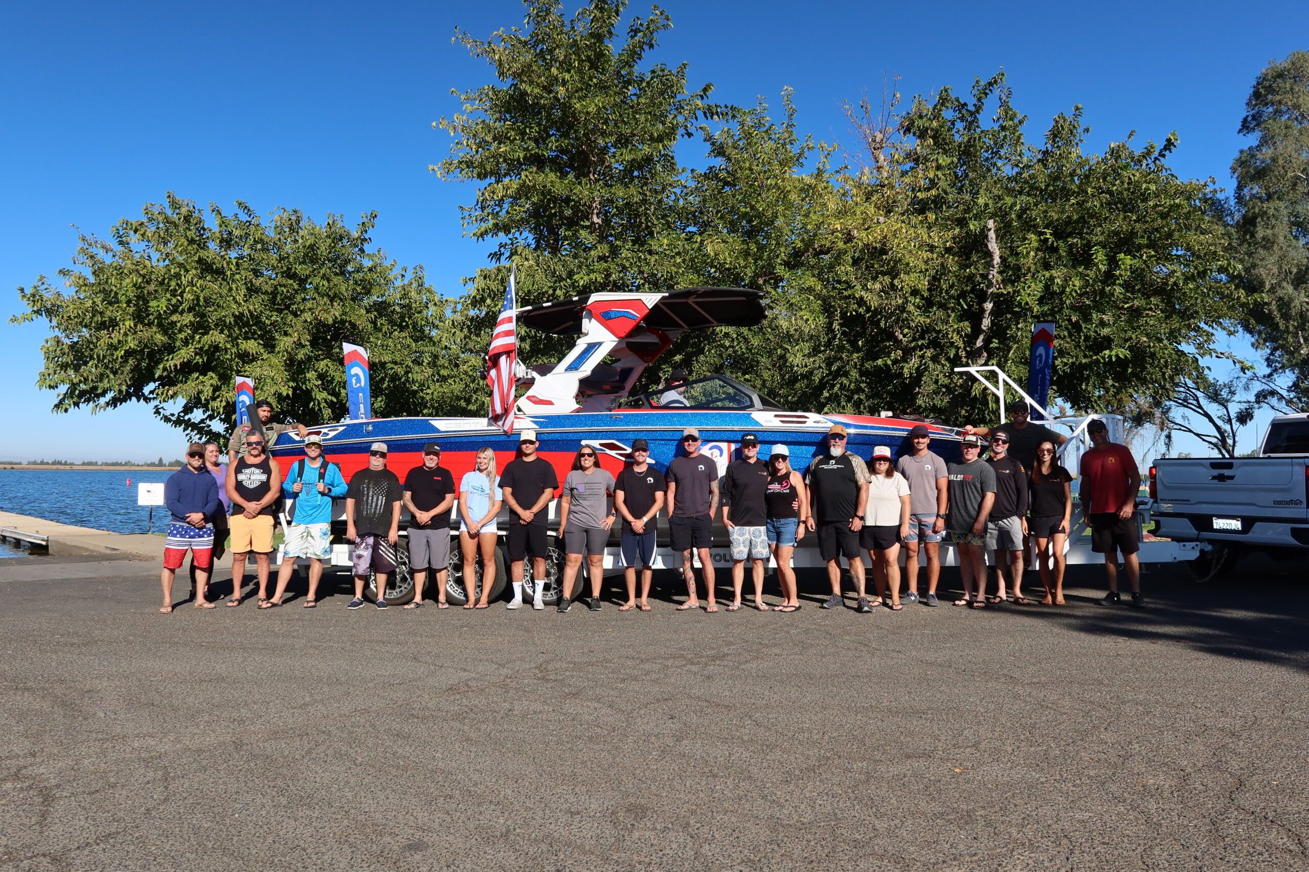 A group of people stands in a line on pavement near a large boat with flags, parked by trees and water on a sunny day.