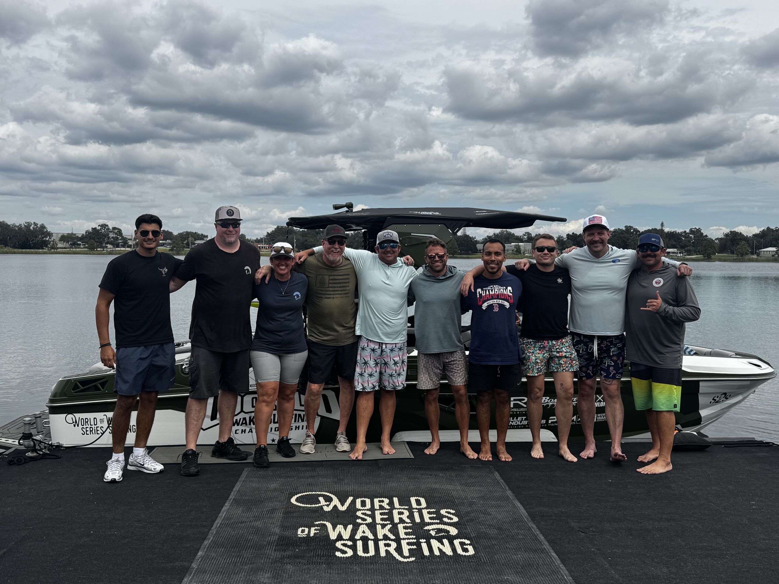 Ten people stand side by side on a dock in front of a boat, smiling, with a “World Series of Wake Surfing” logo on the dock surface and a lake in the background under a cloudy sky.