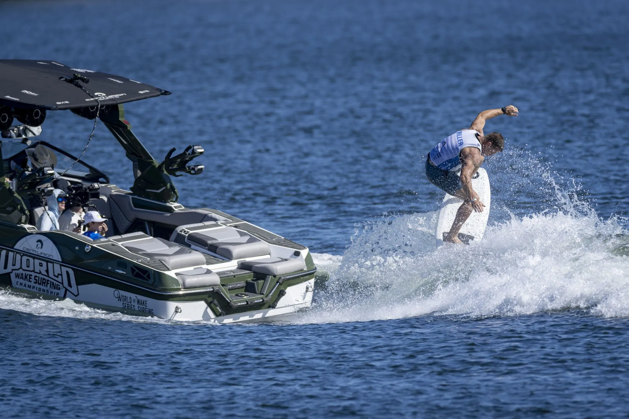 A person wake surfs behind a boat labeled “World Wake Surfing” on a sunny day, with two people seated inside the boat.
