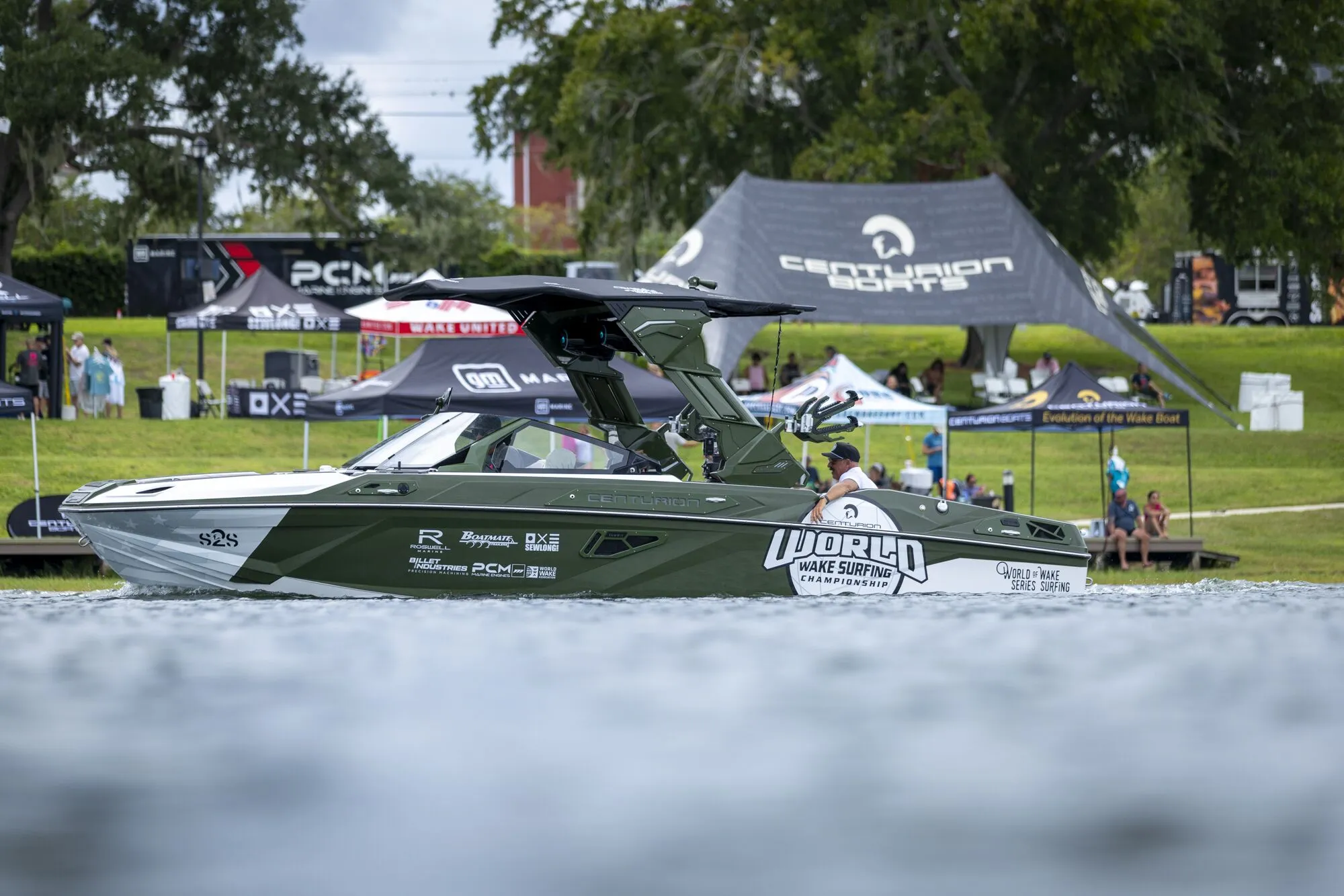 A green and white wakeboarding boat with “World Wake Surfing” decals floats on water near event tents and spectators on a grassy bank.