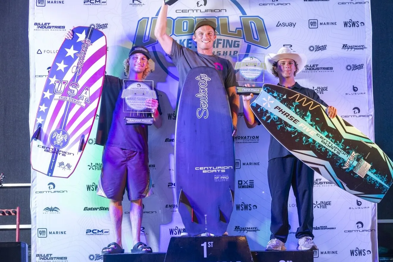 Three athletes stand on a winners’ podium holding trophies and wakeboards, with a banner reading “World Wake Surfing Championship” in the background.