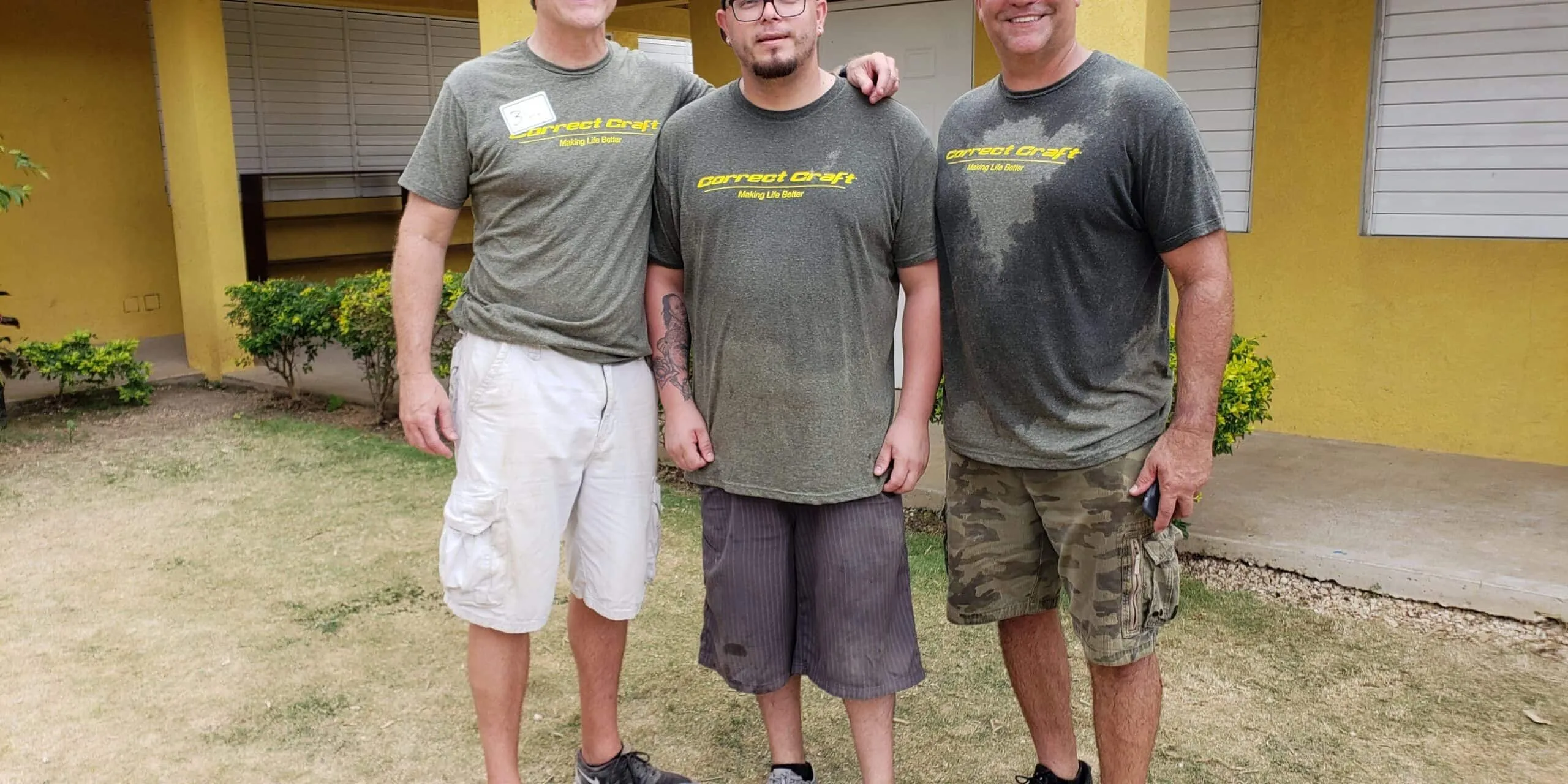 Three men wearing matching green Correct Craft t-shirts standing in front of a yellow building.