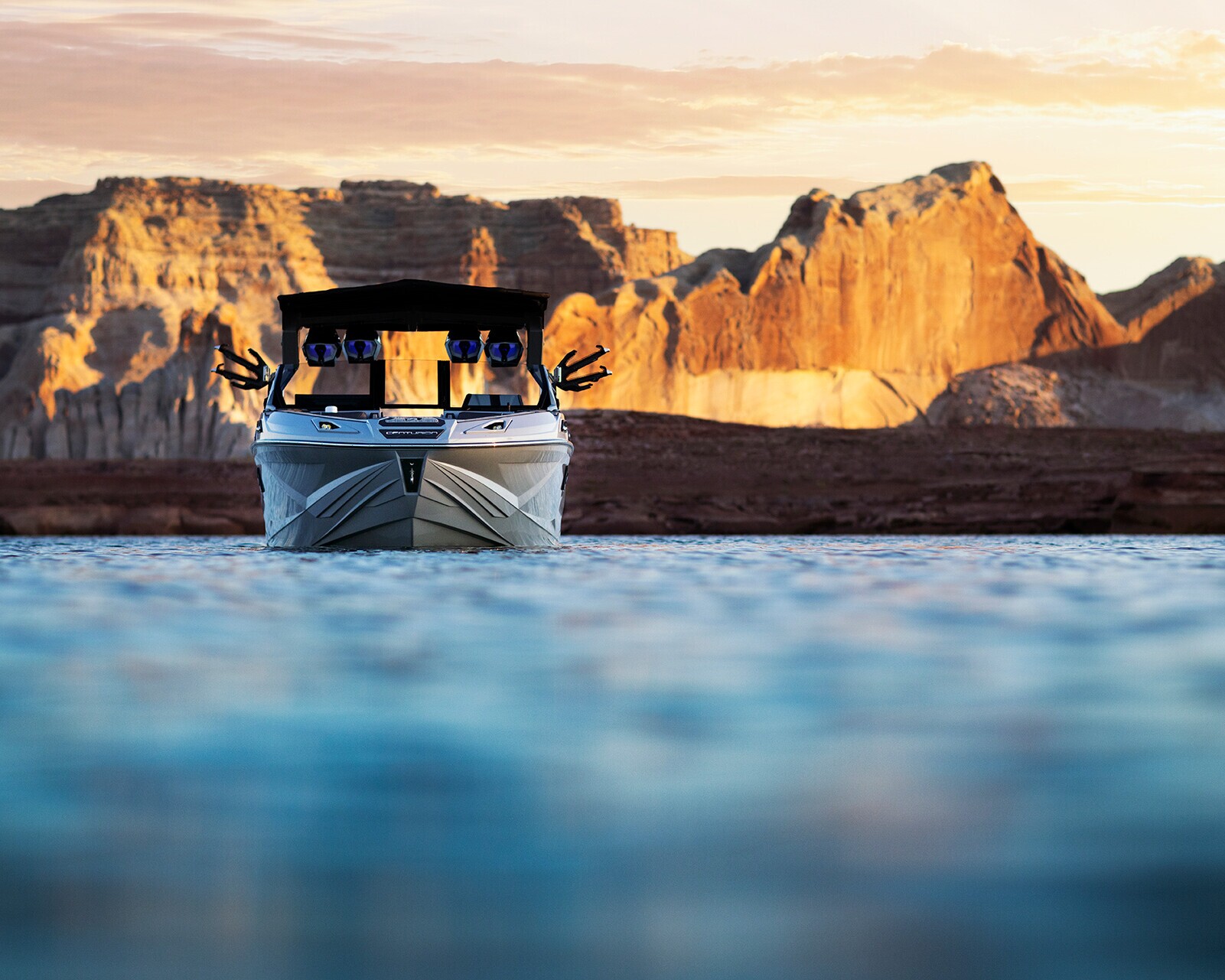 A white Centurion Ri230 glides gently on calm blue water beneath a black canopy, with rocky cliffs and a golden sunset painting the background.