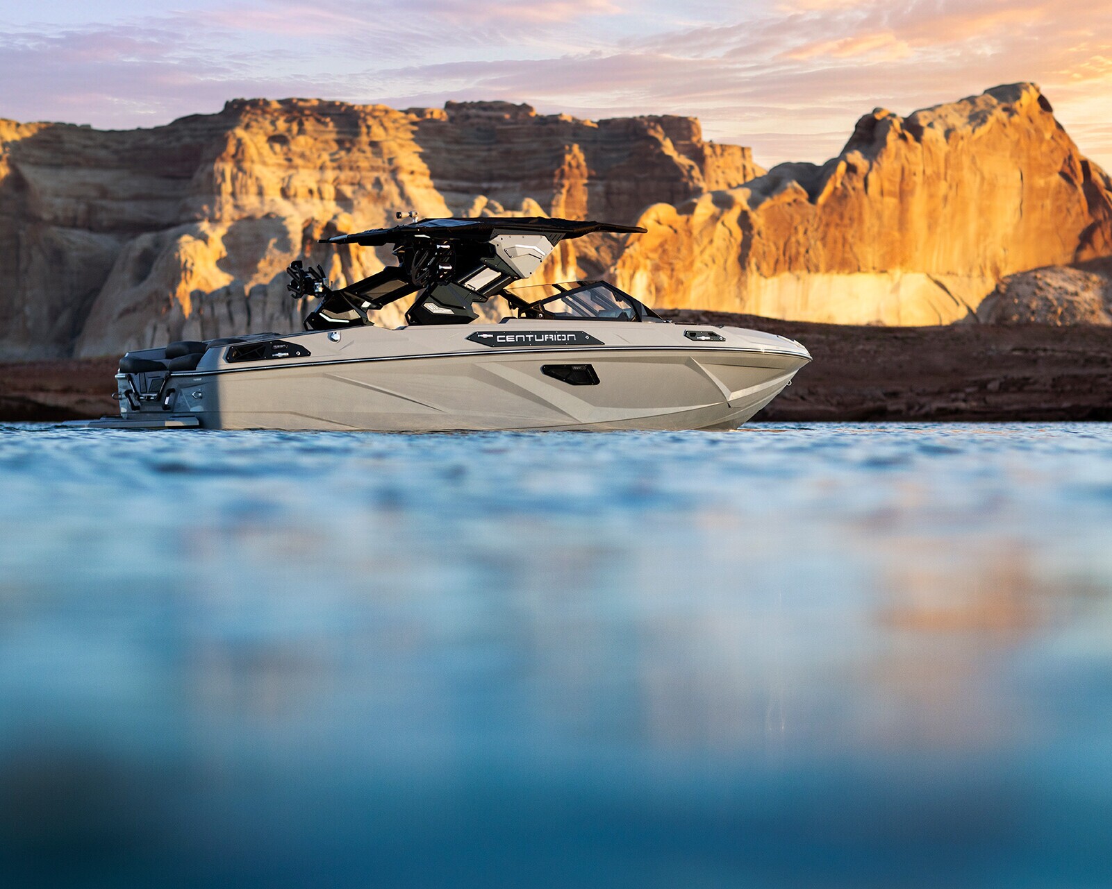 A Centurion Ri230 Wake Boat floats on calm blue water with large rocky cliffs in the background under a partly cloudy sky at sunset.