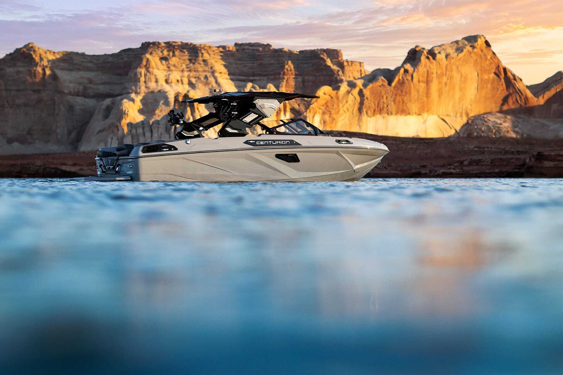 A Centurion Ri230 Wake Boat floats on calm blue water with large rocky cliffs in the background under a partly cloudy sky at sunset.