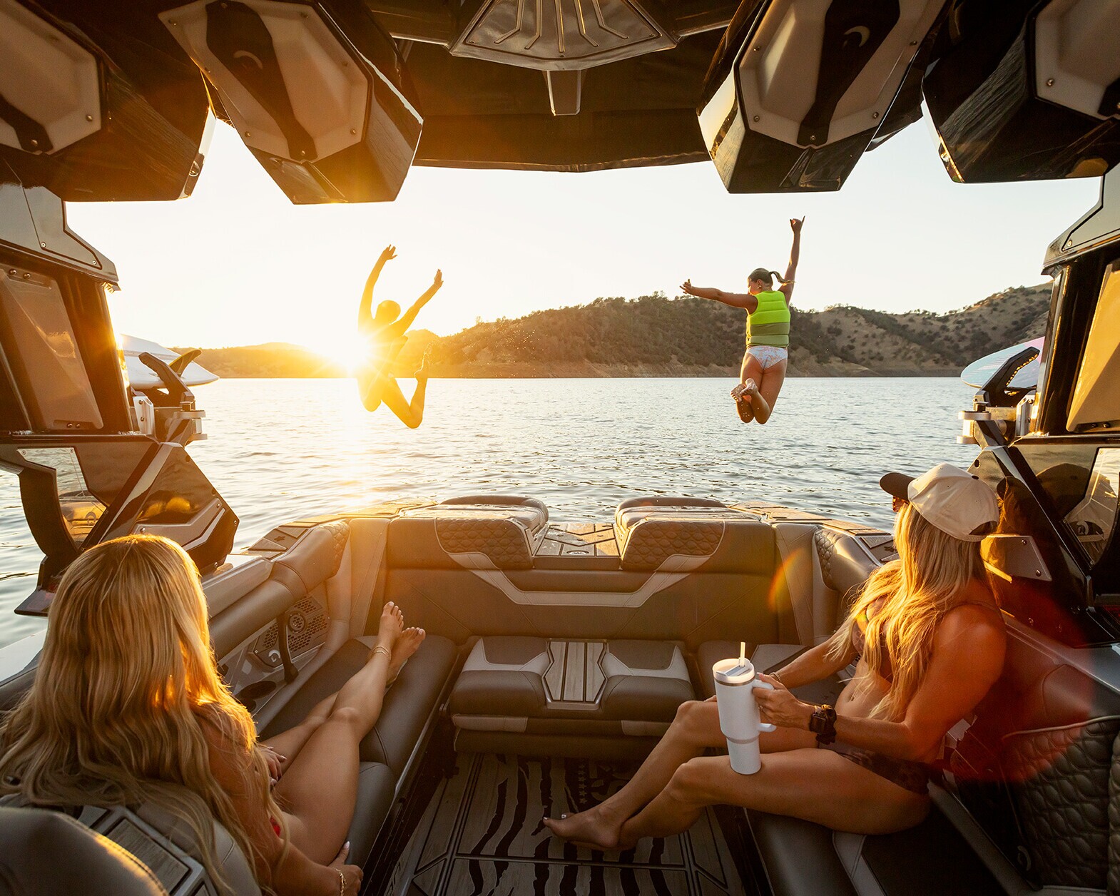 Two people jump off a Centurion Ri230 wake boat into a lake at sunset while two others sit and watch from inside the boat, surrounded by hills.