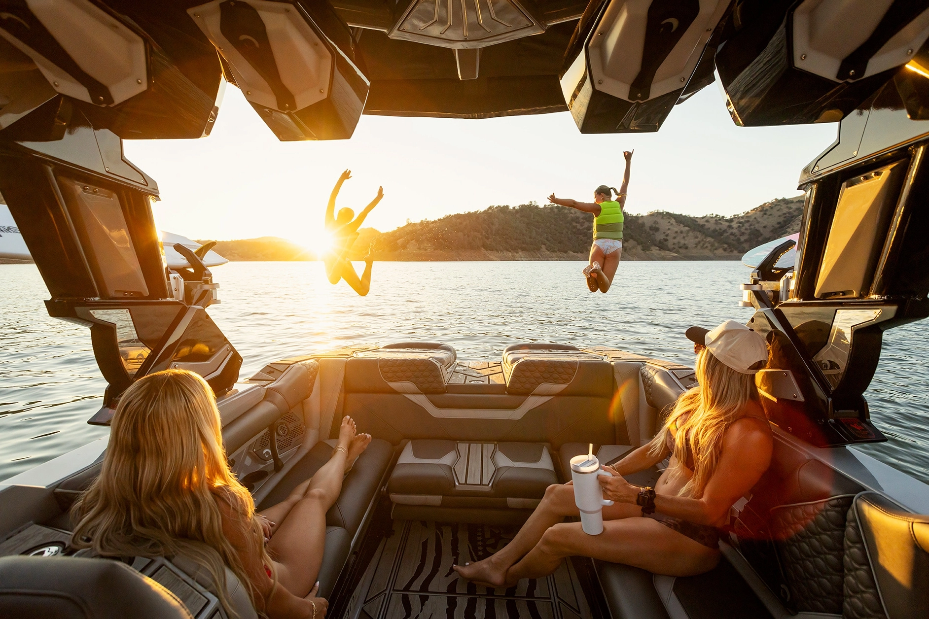 Two people jump off a Centurion Ri230 wake boat into a lake at sunset while two others sit and watch from inside the boat, surrounded by hills.