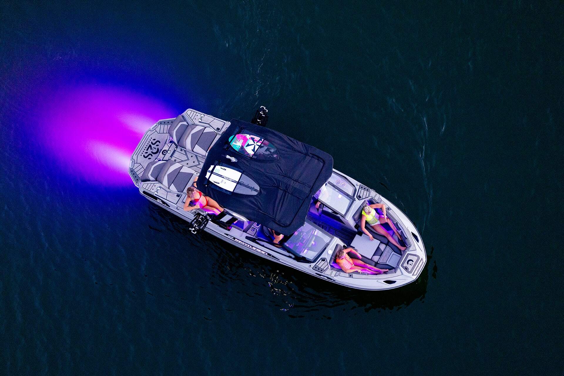 Overhead view of a Centurion Ri230 Wake Boat with four people on board, traveling on dark water, with purple light glowing from the rear.