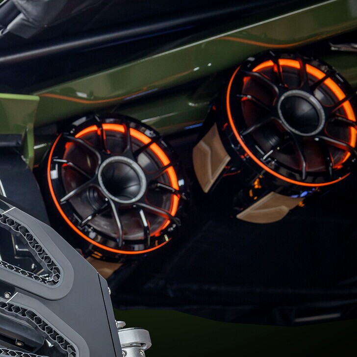 Close-up of two circular speakers with orange LED lights mounted on a dark green vehicle, with part of a metallic mechanical component visible in the foreground.
