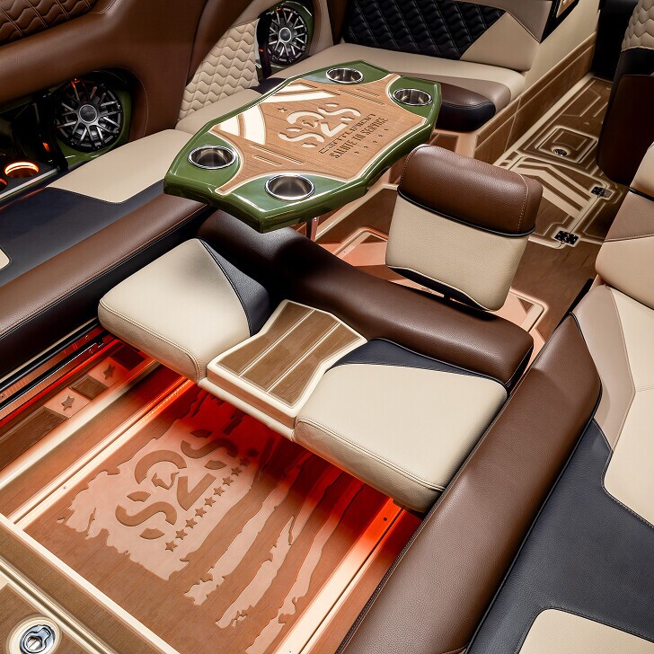 Luxury vehicle interior featuring custom leather seats, a wood and leather table with cup holders, and detailed flooring with an American flag design, all in brown, beige, and black tones.