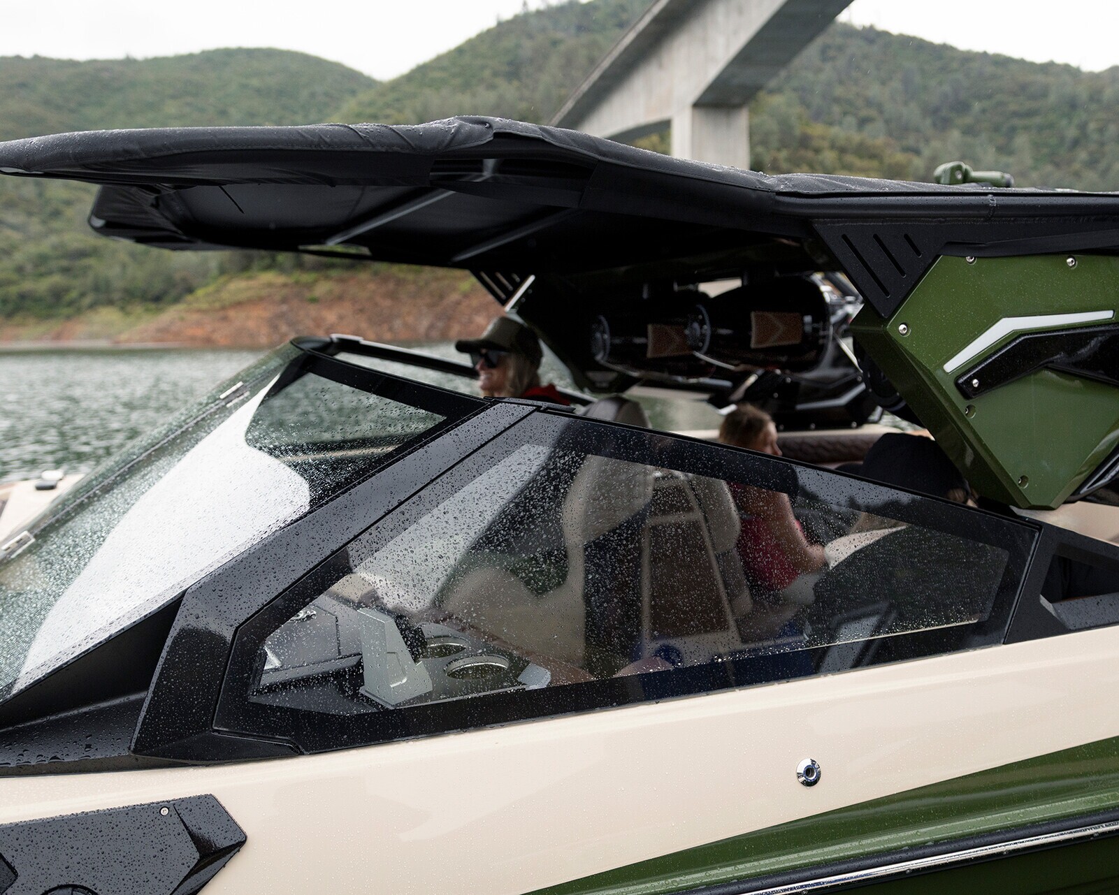 A close-up of a Centurion Ri245 motorboat with people seated inside, docked on a lake with a bridge and tree-covered hills in the background. The boat features a dark canopy and green accents.