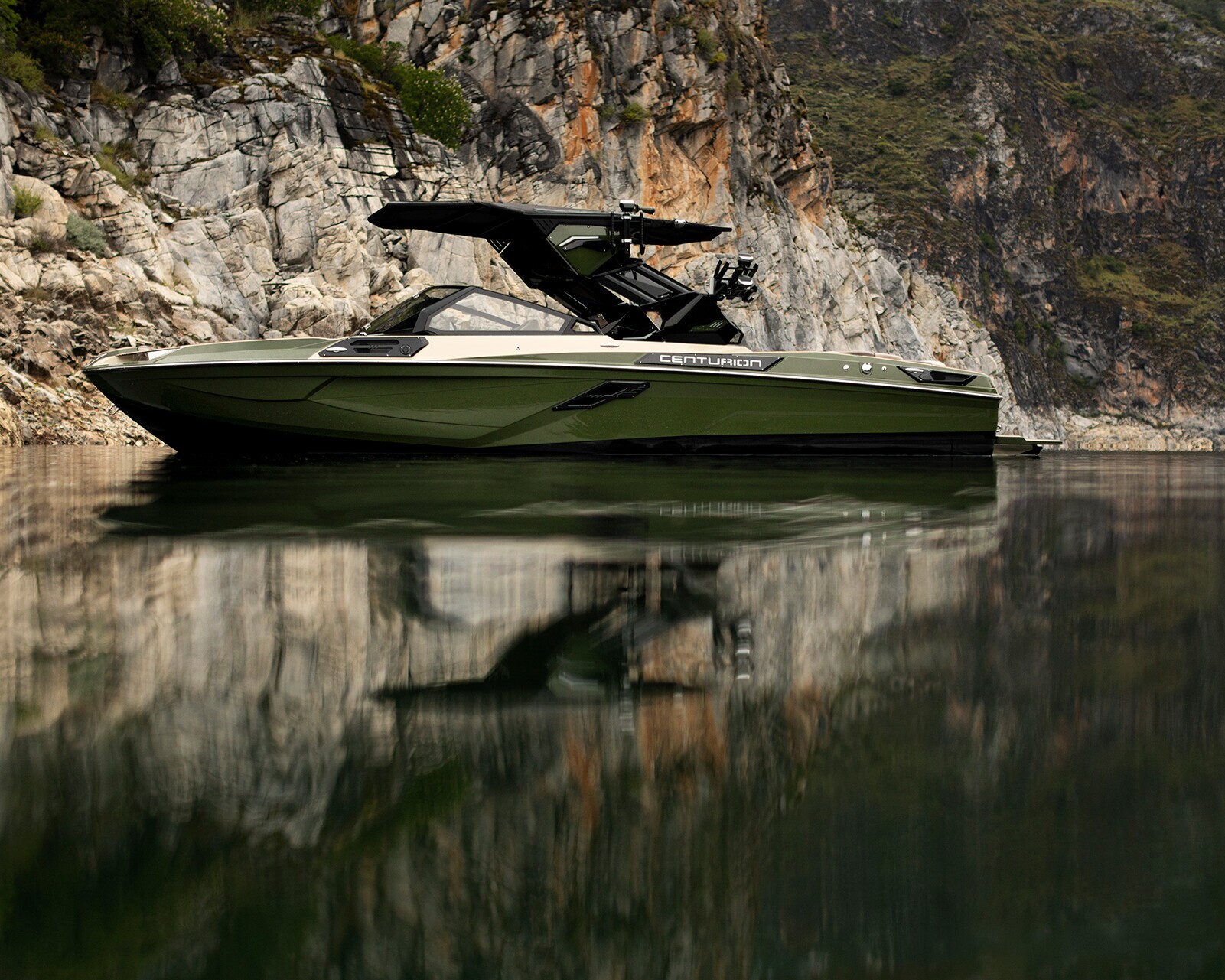 A green Centurion Ri245 is anchored near rocky cliffs, its sleek hull reflecting on the calm water of a lake or river.