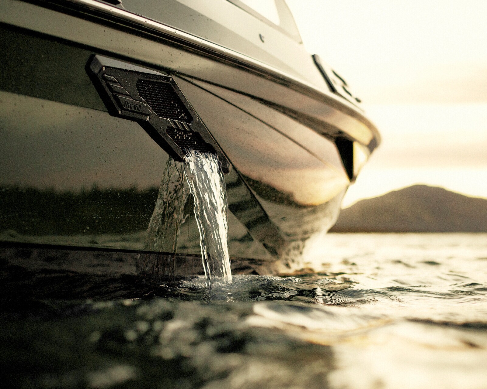 Close-up of water draining from a vent on the side of a Centurion Ri245 surf boat floating on a lake at sunset, with mountains visible in the background.