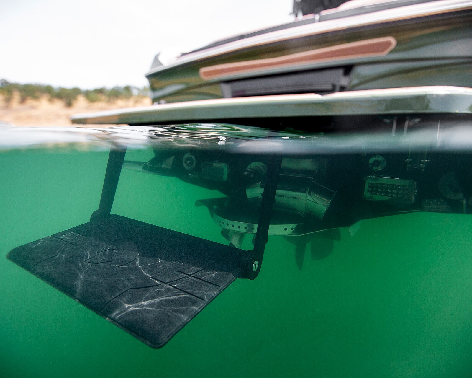 An underwater view of a Centurion Ri245 Surf Boat’s wakeboard surf tab device extended below the hull, with waterline and shoreline visible in the background.