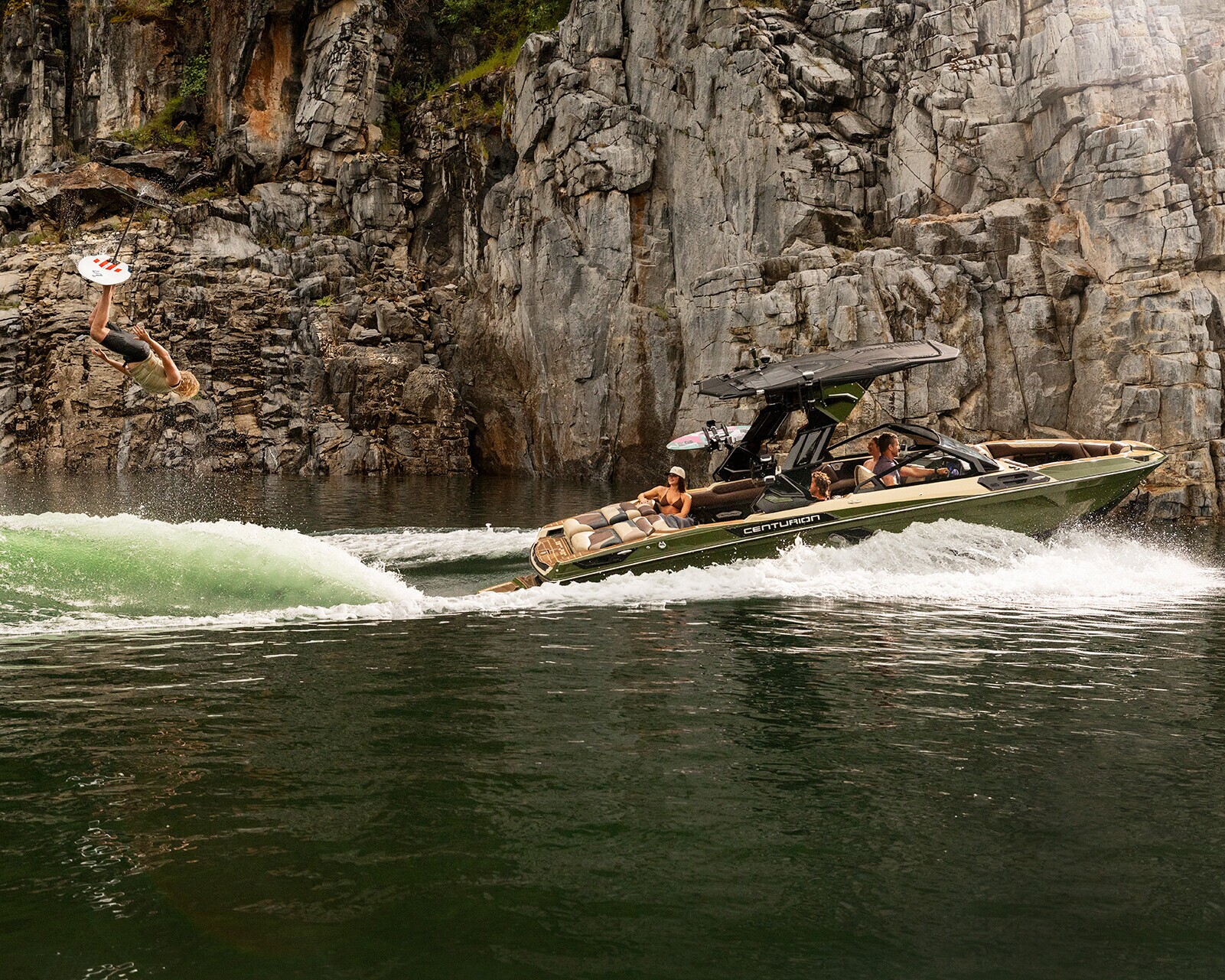 A person wakesurfs behind a Centurion Ri245 near rocky cliffs, with several people watching from the boat on a calm body of water.