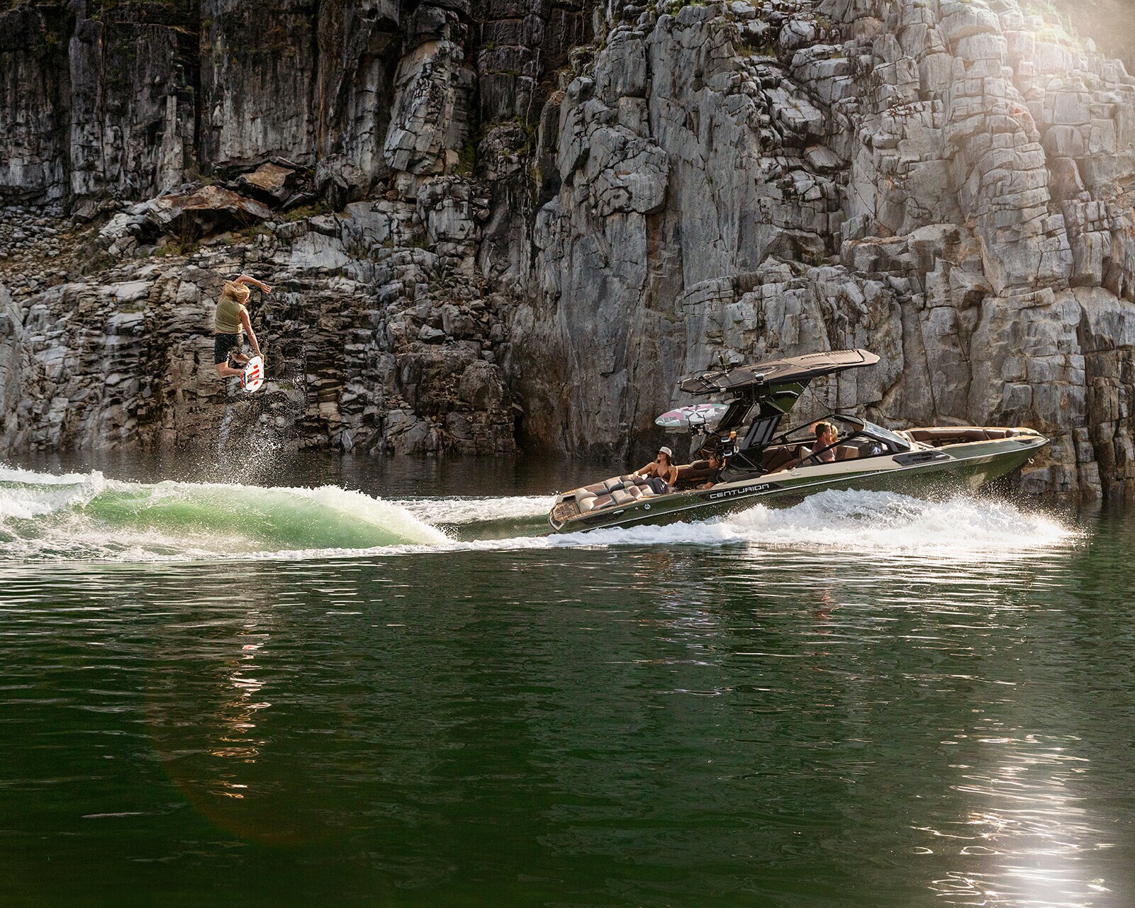 A person is wakeboarding behind a Centurion Ri245 on a green lake, with high rocky cliffs in the background and sunlight reflecting on the water.