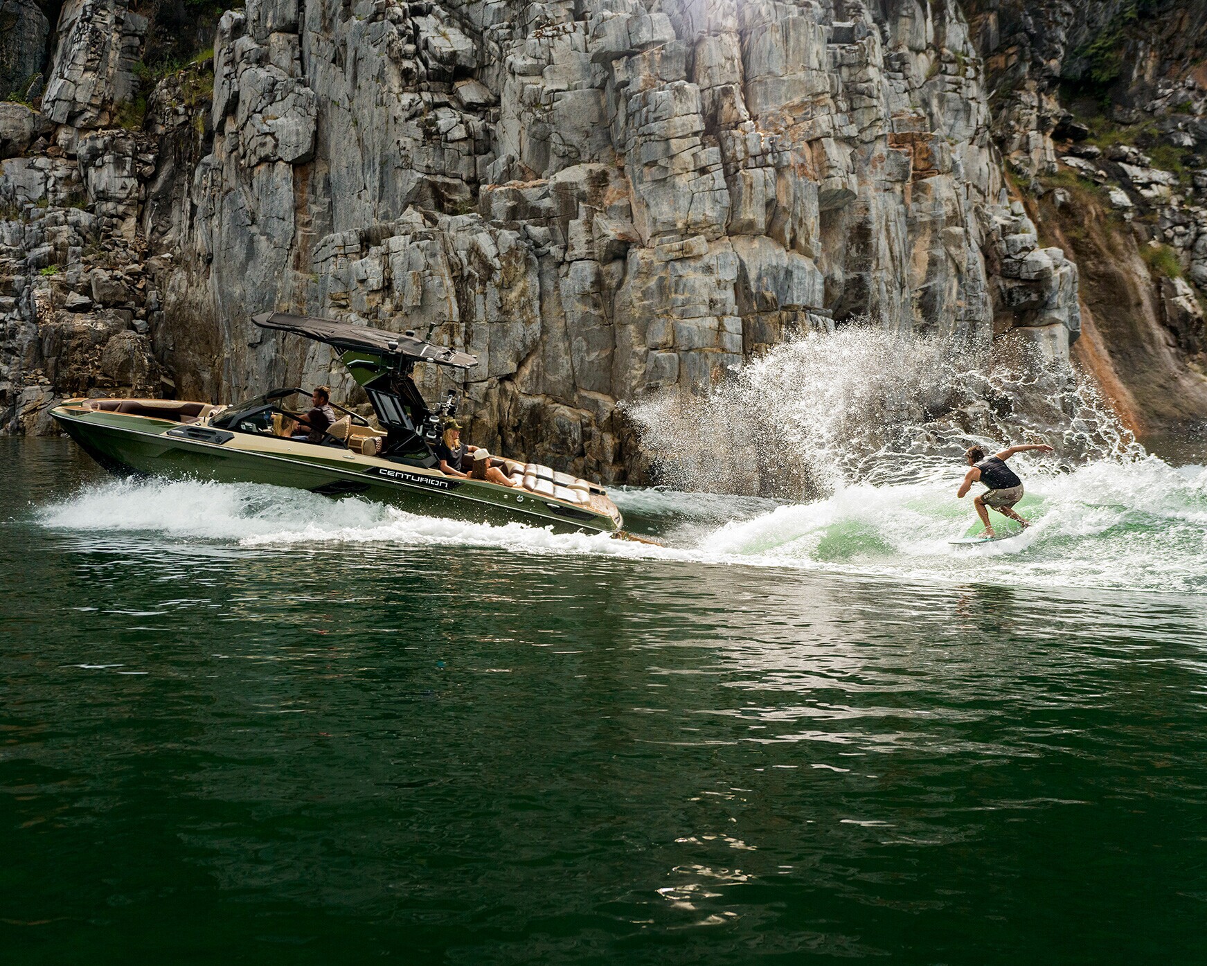 A person is wake surfing on a wave behind a Centurion Ri245 in a green body of water, with tall rocky cliffs in the background.