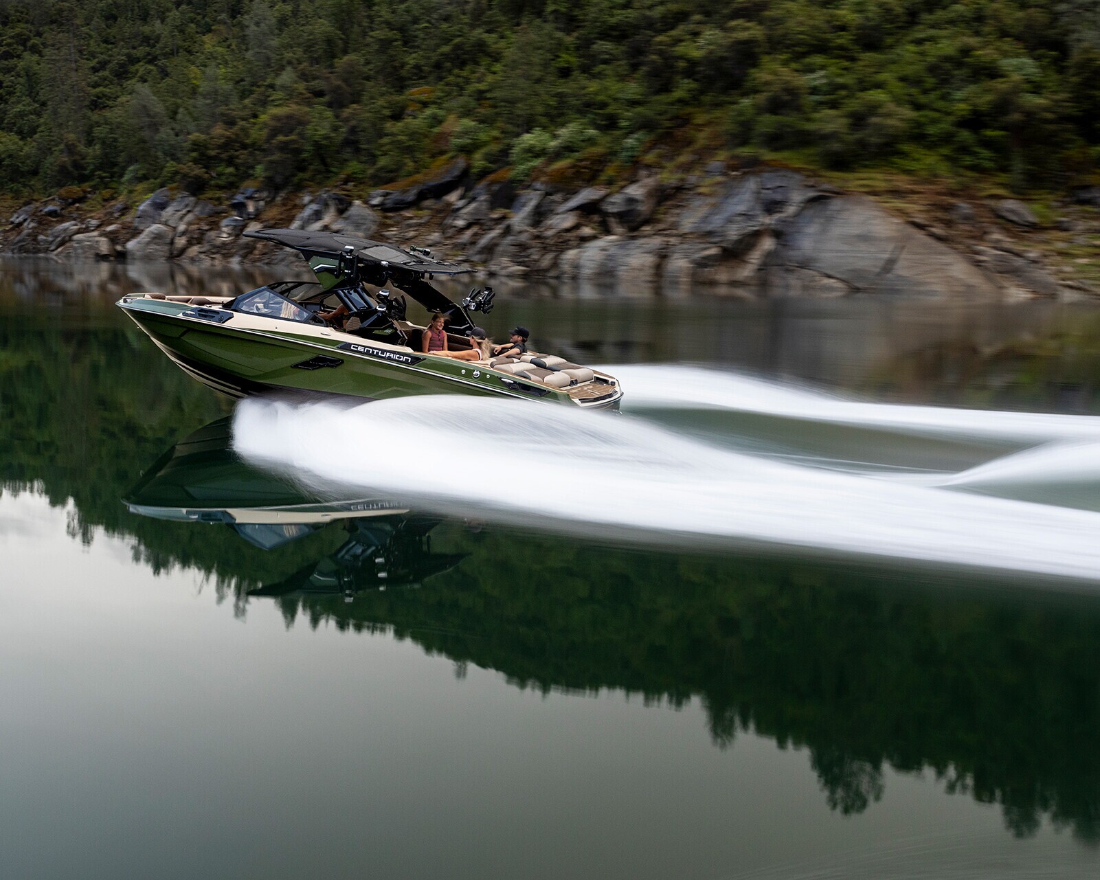 A Centurion Ri245 powerfully glides across a calm lake, leaving a trail of white wake behind, with forested hills and rocks in the background.
