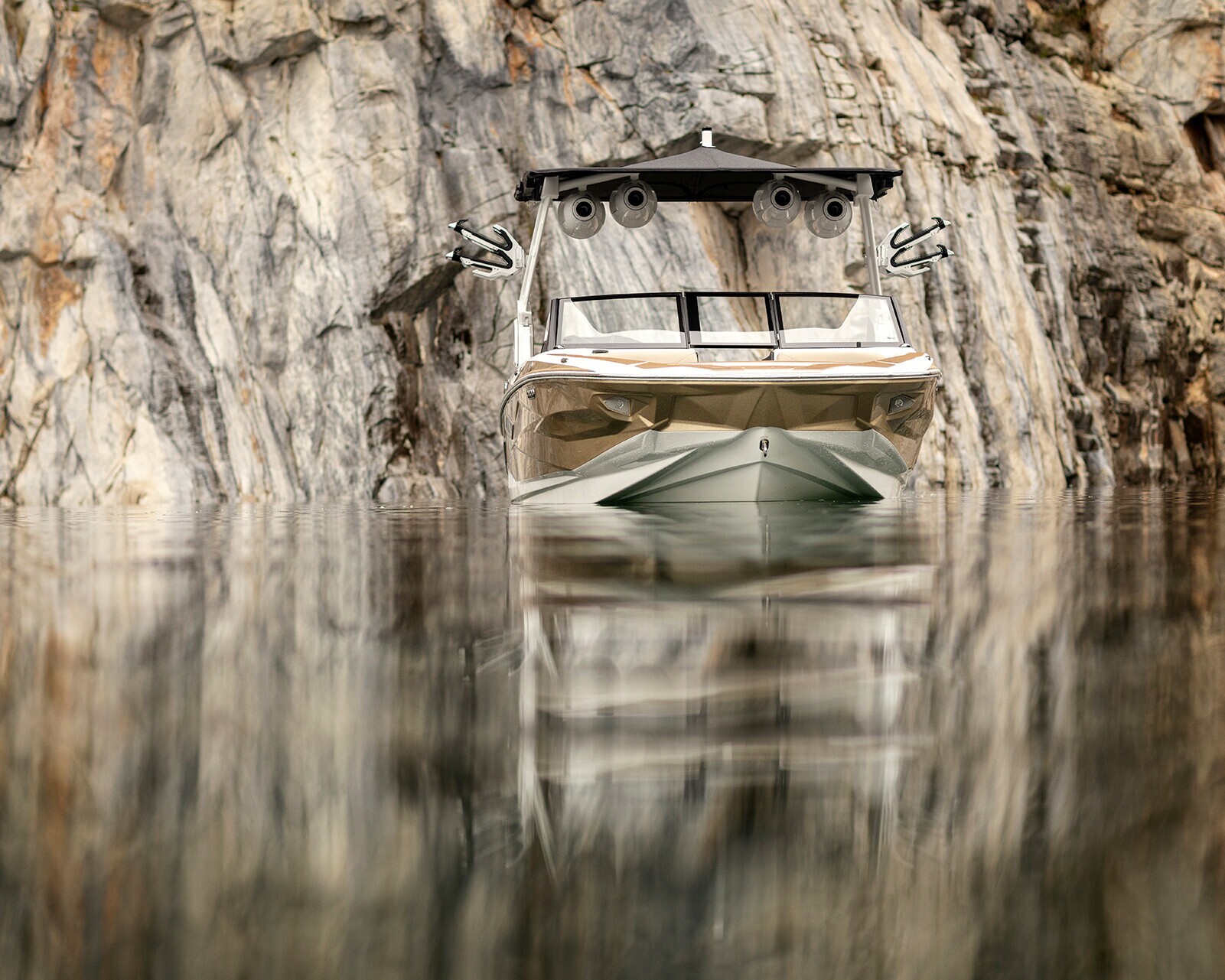 A modern Centurion Nv213 surf boat is stationary on calm water near a steep, rocky cliff. The water reflects the sleek boat and the rugged rock face.