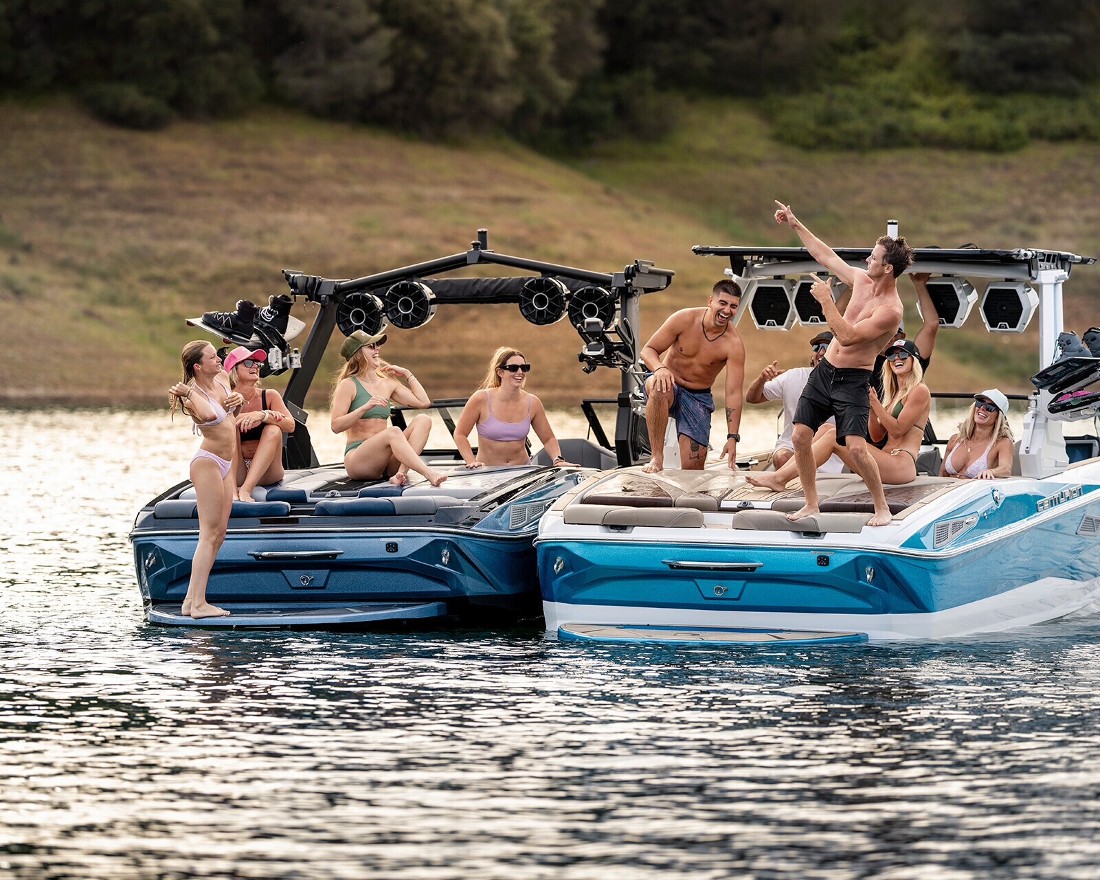 Two motorboats, including a Centurion Nv233, are side by side on a lake. Several people in swimwear relax, chat, and take photos on the Nv233 Wake Boat, while one person stands and points toward the sky.