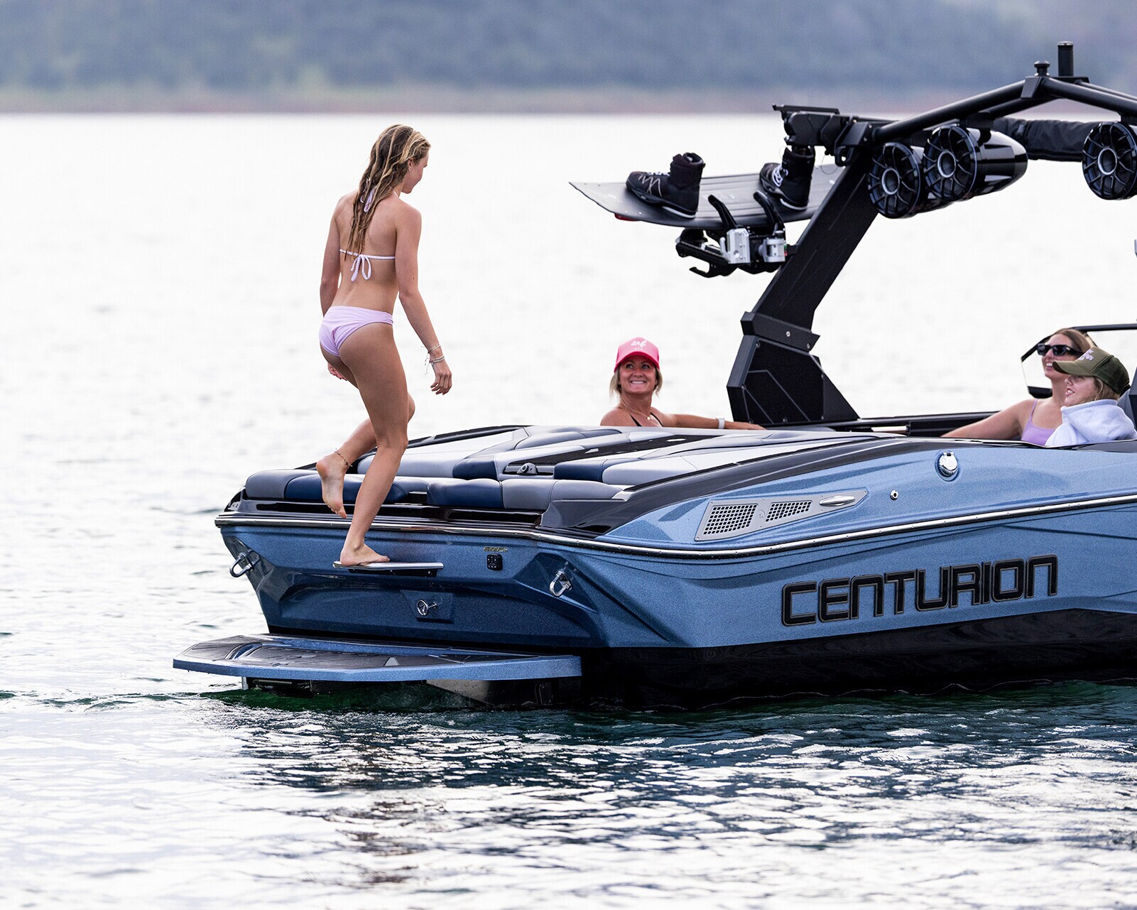 A woman in a pink bikini steps onto a blue Centurion Nv233 wake boat where two people are seated, one wearing a pink cap, on a calm body of water.