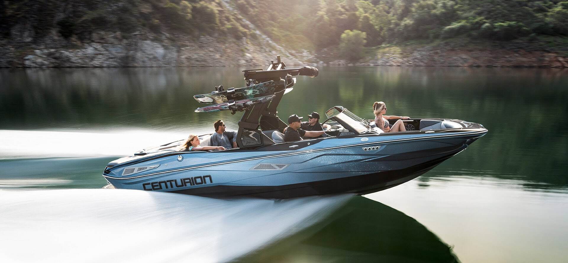 A blue Centurion Nv233 speedboat with four people onboard moves quickly across a calm lake, surrounded by trees and a rocky shoreline.